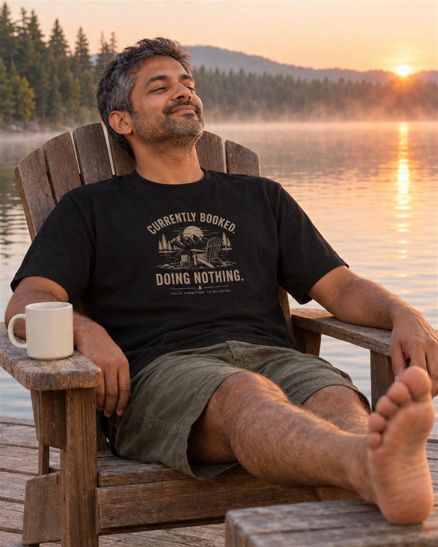 Man relaxing on a lakeside chair in a Currently Booked tee
